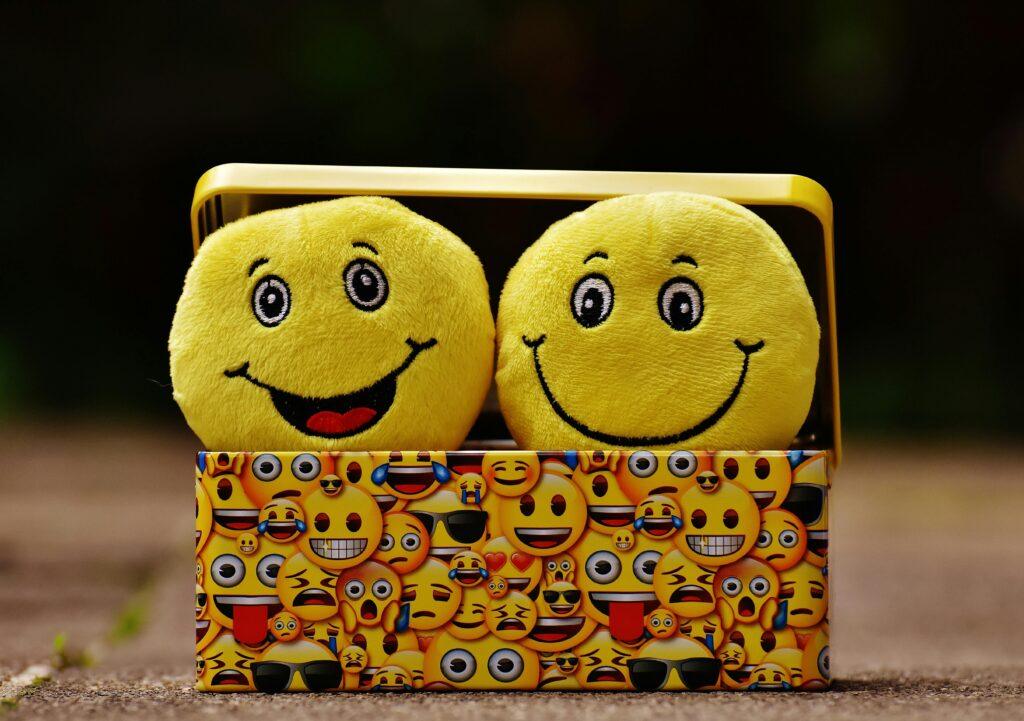 Two yellow smiley plush toys in an emoji-themed box, expressing fun and happiness.